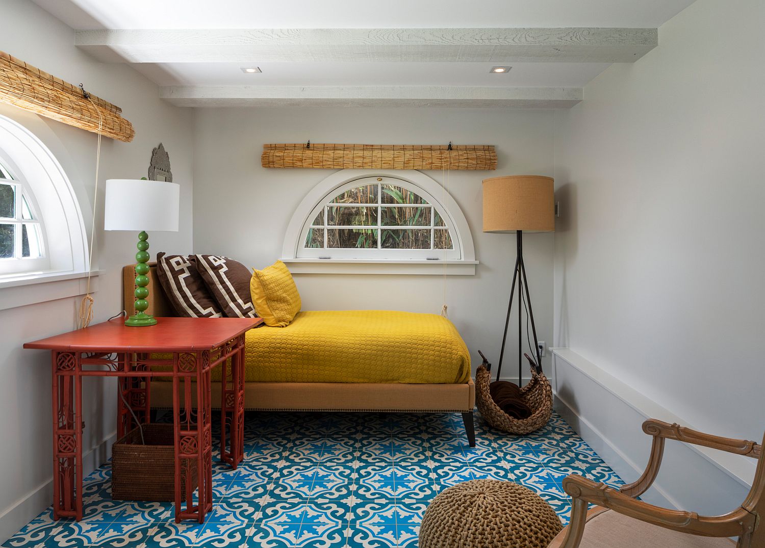 Hình ảnh phòng ngủ nhỏ phong cách Bắc Âu với tường và trần sơn trắng, ga gối màu vàng chanh, bàn đỏ bên cạnh, rèm cửa bằng tre che cửa sổ kính bán nguyệt, sàn lát gạch bông xanh dương.