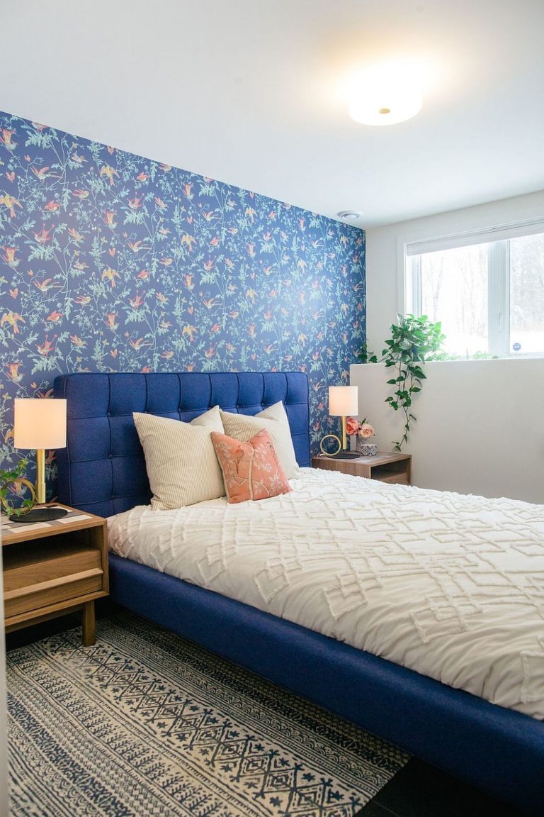 Hình ảnh phòng ngủ với nệm màu trắng, giường xanh, tường đầu giường dán giấy họa tiết hoa lá, chim muông, cửa sổ kính ngập tràn ánh sáng.