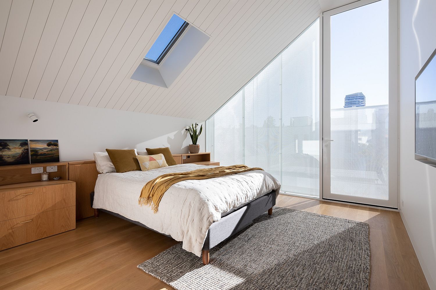 Hình ảnh phòng ngủ nhỏ trên tầng áp mái với tường và trần màu trắng tinh khôi, sàn nhà lát gỗ tạo cảm giác ấm áp, khung cửa sổ kính lớn đón sáng tự nhiên.