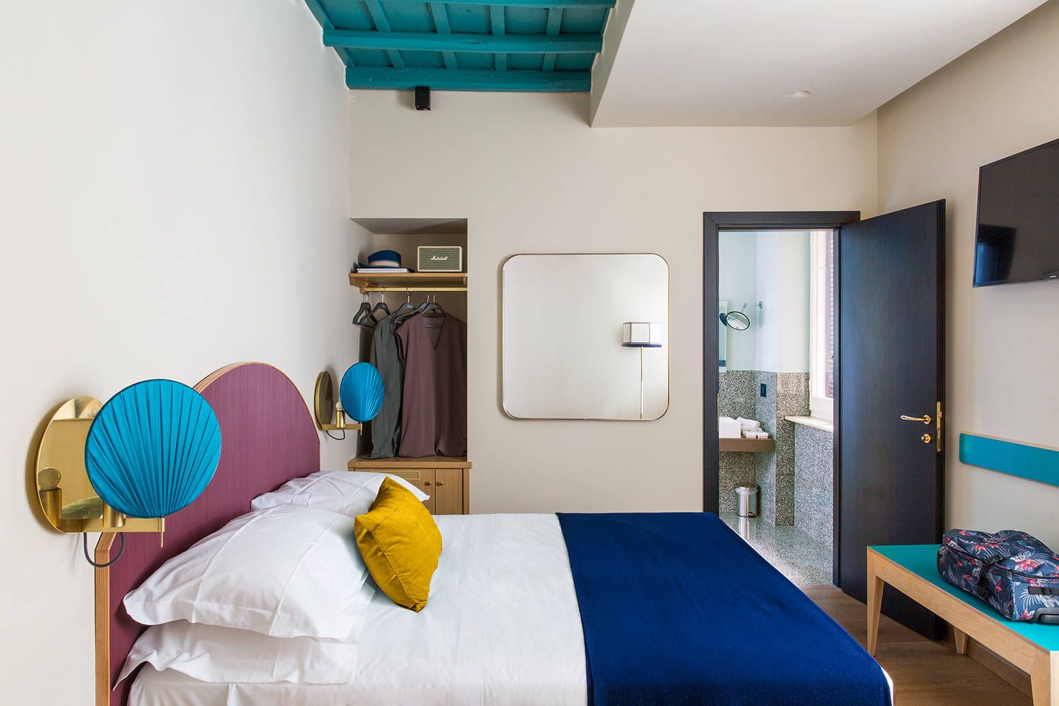 Hình ảnh phòng ngủ nhỏ với ga gối màu trắng, chăn mỏng xanh dương, đầu giường sơn tím oải hương, bàn cuối giường xanhngocj lam, cạnh đó là tủ quần áo âm tường.