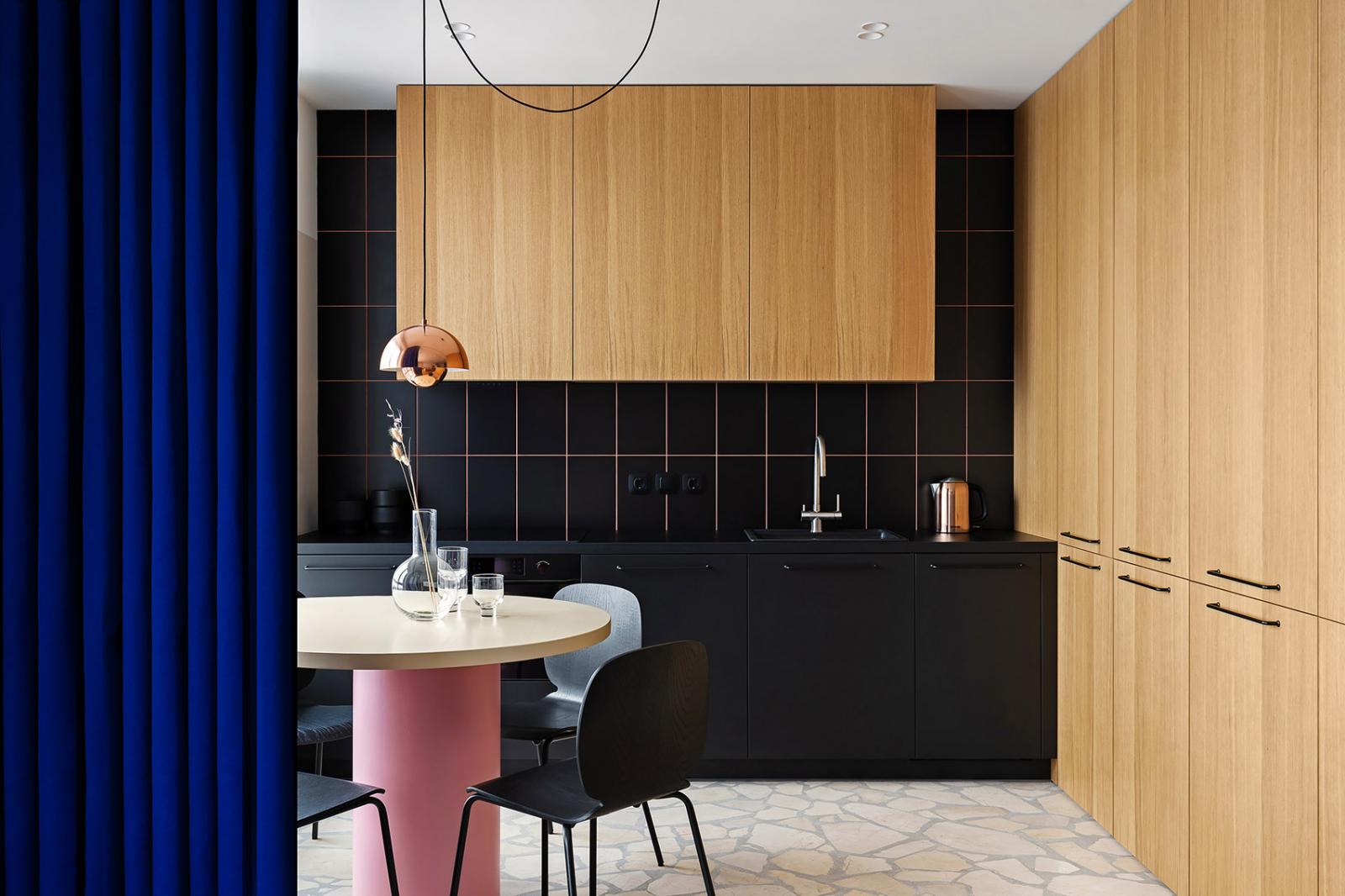 Hình ảnh phòng bếp với tường ốp gạch màu đen, cùng tông với tủ dưới, tủ trên bằng gỗ màu sáng, cạnh đó là bàn ăn hình tròn màu hồng và rèm cửa màu xanh coban