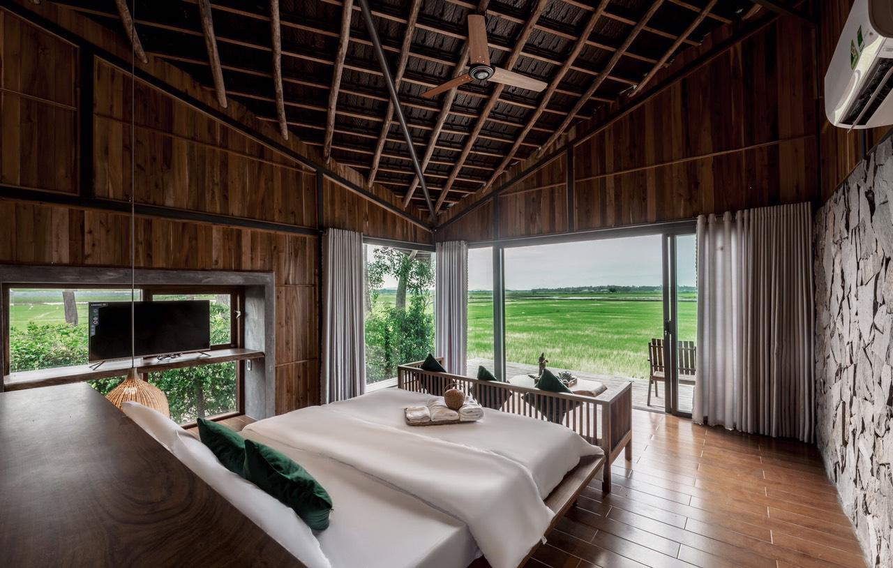 Hình ảnh phòng ngủ với ga gối màu trắng, tường và trần ốp gỗ, nhìn thẳng ra ruộng lúa