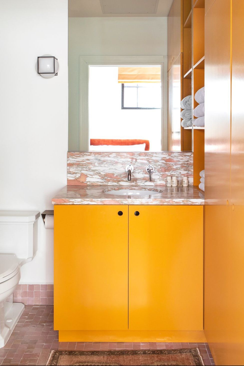 Hình ảnh phòng tắm nhỏ với tủ màu cam nhạt cùng tông với bức tường liền kề, sàn lát gạch hồng, thảm thổ cẩm