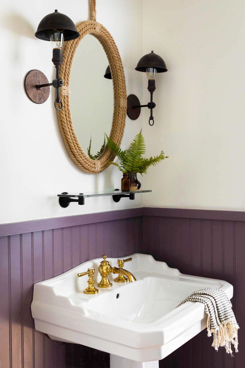 Hình ảnh phòng tắm nhỏ với tường phía dưới sơn màu tím nhạt, phía trên là màu trắng, khung gương dây thừng bắt mắt