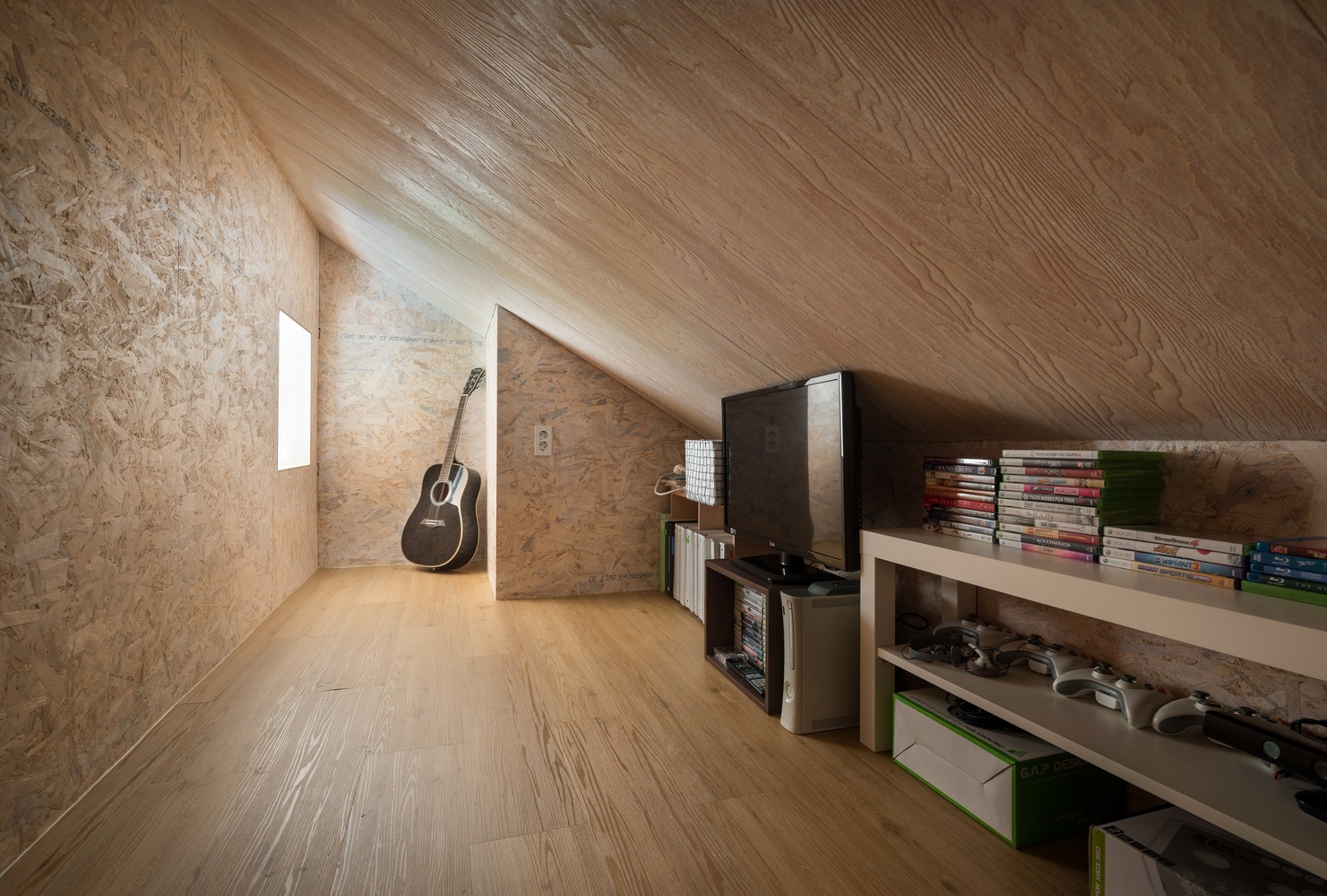 Hình ảnh phòng làm việc trên tầng áp mái với trần và tường ốp gỗ, kệ sách mở, tivi, đàn ghita ở góc
