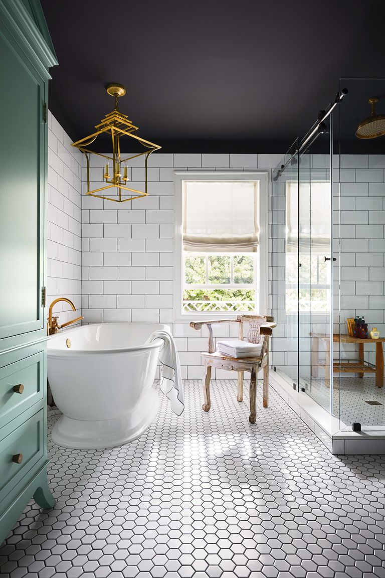 Hình ảnh một phòng tắm thoáng sáng với trần sơn đen, tường và sàn ốp gạch men màu trắng, đèn trang trí mạ đồng sáng bóng, bồn tắm nằm cạnh buồng tắm cạnh, giữa là ghế ngồi, cạnh đó là tủ đựng đồ màu xanh dương