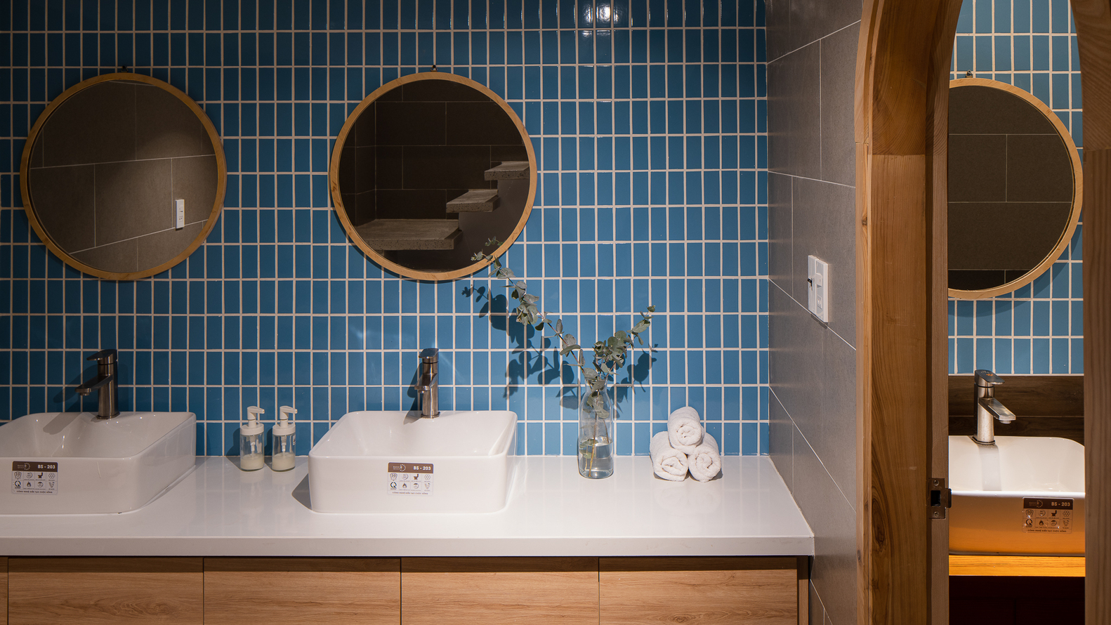 Hình ảnh một góc phòng tắm với tường ốp gạch màu xanh dương, gương tròn đôi, bồn rửa đôi