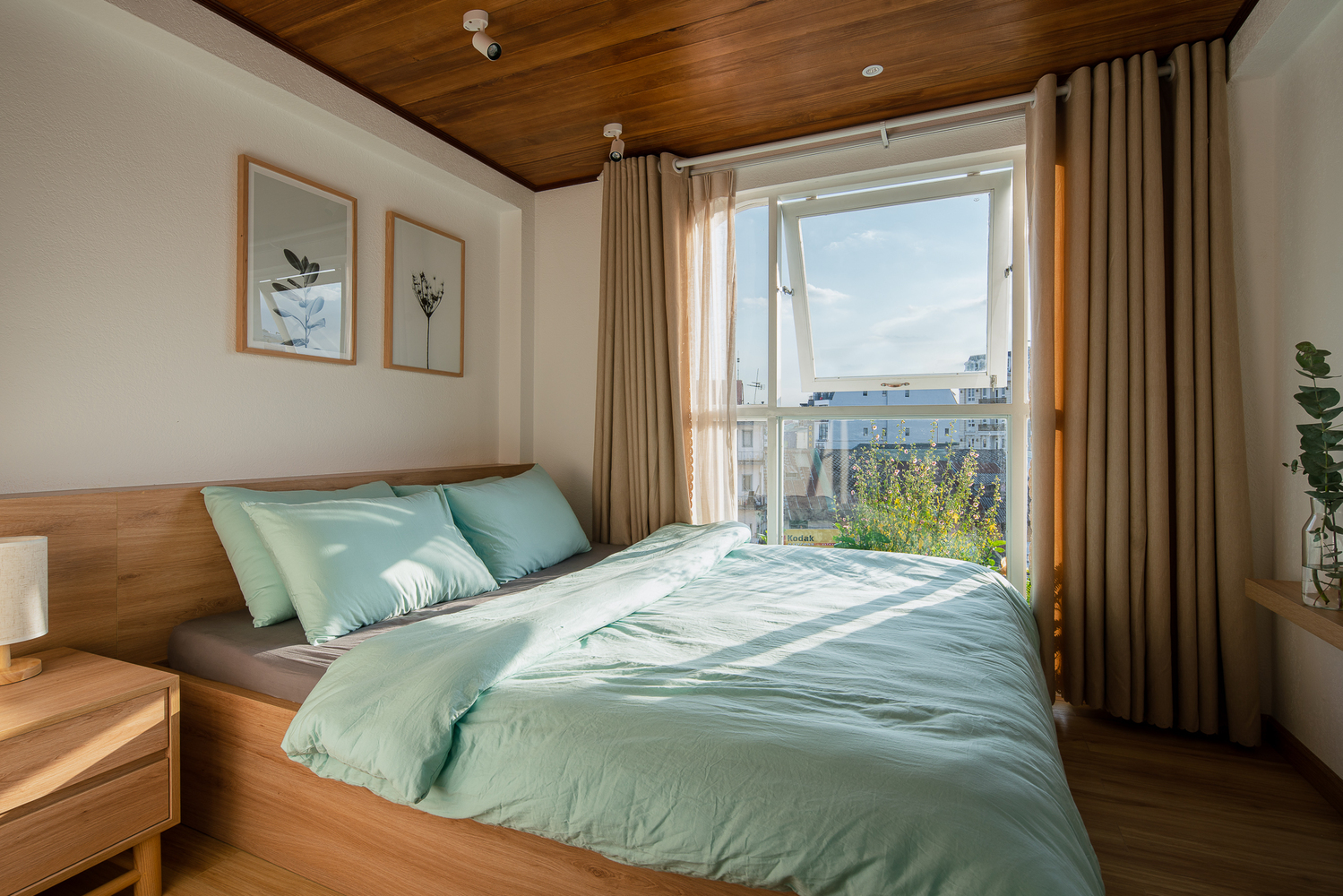 Hình ảnh phòng ngủ rộng thoáng với ga gối màu xanh ngọc lam dịu mát, cạnh đó cửa sổ kính lớn, tranh treo đầu giường