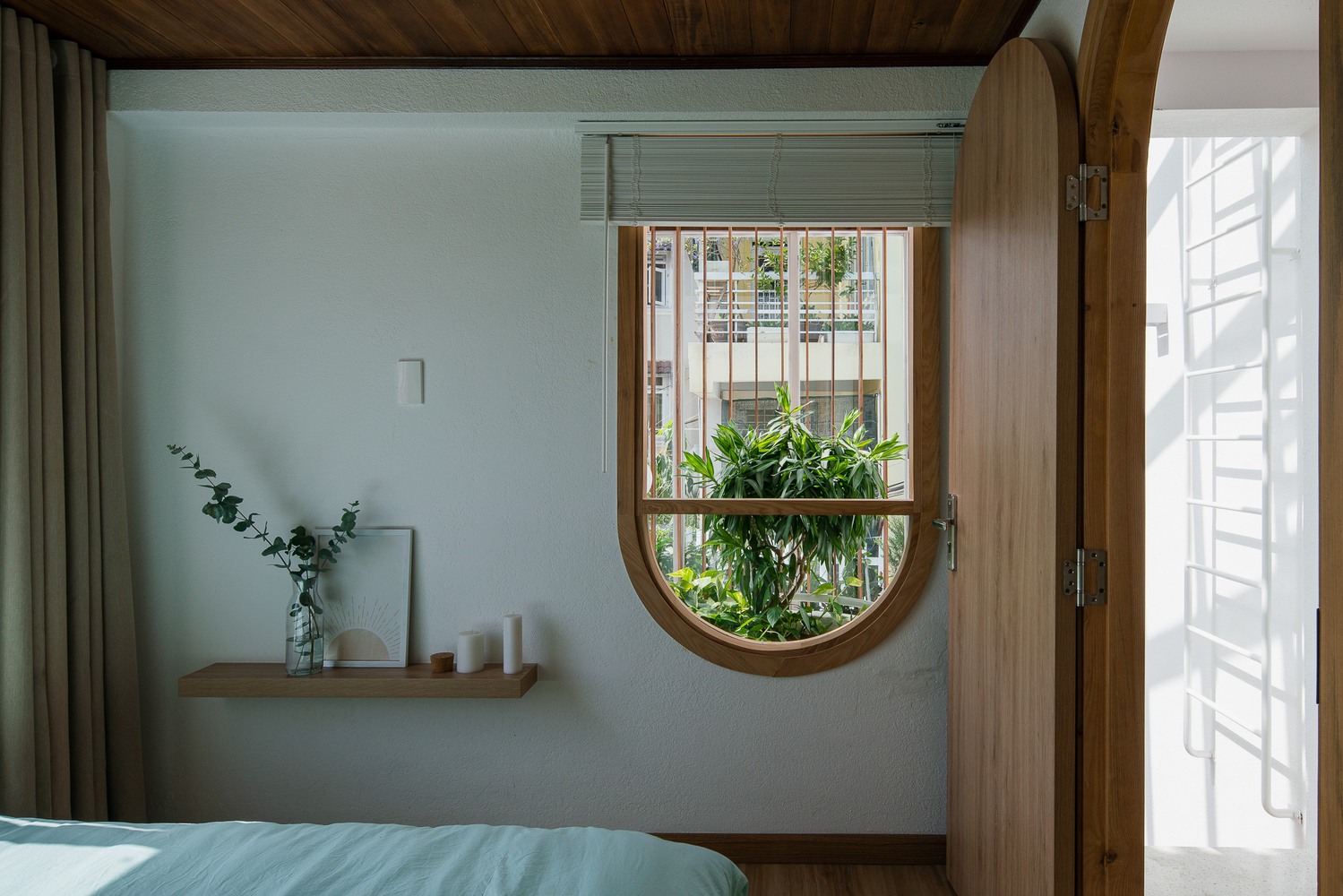 Hình ảnh một góc phòng ngủ với kệ mở gắn tường đặt bình cây, cửa sổ vòm mở ra khoảng xanh bên ngoài