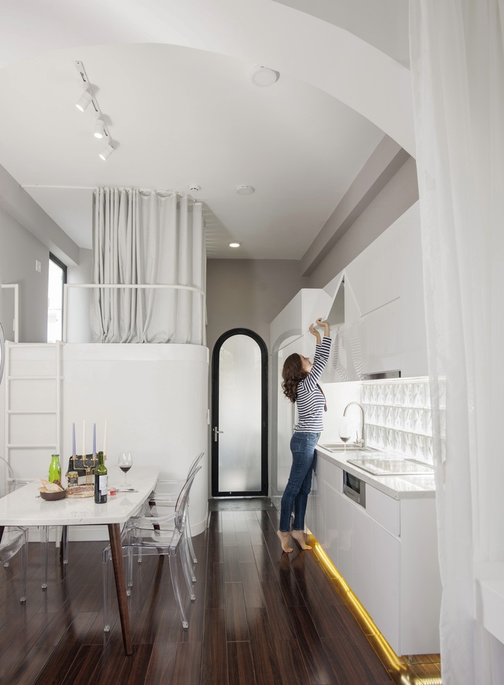 Hình ảnh phòng bếp ăn phong cách Paris với bàn đá màu trắng, sàn lát gỗ bóng, người phụ nữ đang với tay mở cửa tủ bếp trên