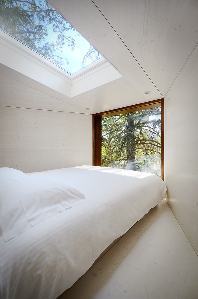 Hình ảnh cận cảnh giường nệm màu trắng, cửa sổ mái trong suốt, bên ngoài nhiều cây xanh