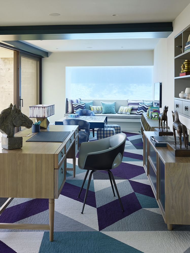 Hình ảnh cận cảnh bàn làm việc bằng gỗ có ngăn kéo, ghế tựa đặt trên thảm trải họa tiết hình học màu xanh, cạnh đó là phòng khách thoáng sáng