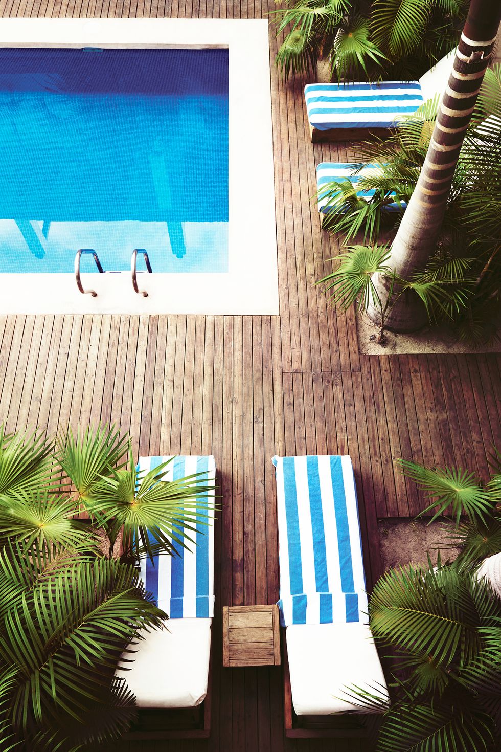 Hình ảnh bể bơi nhìn từ trên cao xuống với nước xanh ngắt, ván sàn gỗ lát xung quanh, ghế tắm nắng kẻ sọc xanh trắng
