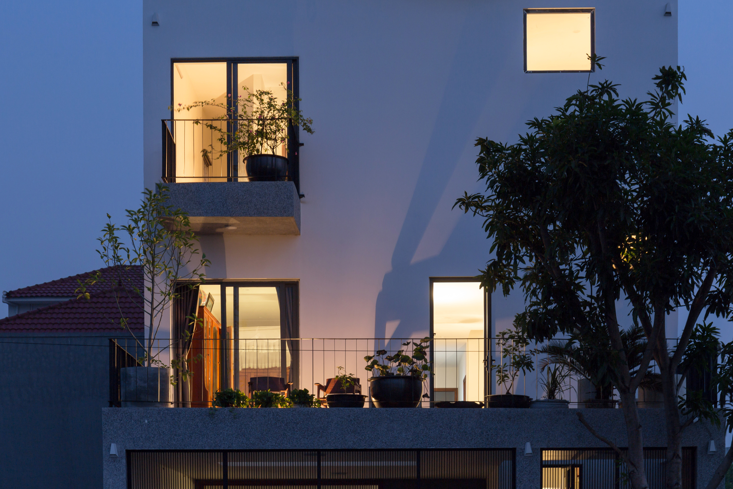 Hình ảnh cận cảnh mặt tiền các tầng trên của nhà phố khi đêm xuống với cửa sổ kính trong suốt, cây xanh trồng ở ban công