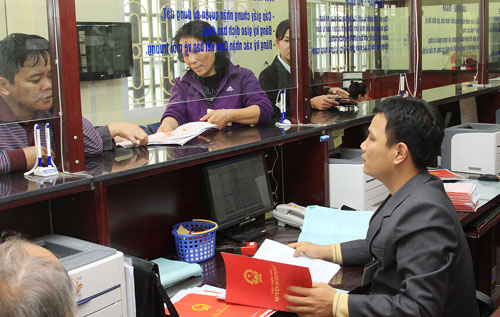 Hình ảnh hai người dân đang đứng đợi làm thủ tục giấy tờ, một nhân viên ngồi trong quầy giao dịch đang cầm sổ đỏ