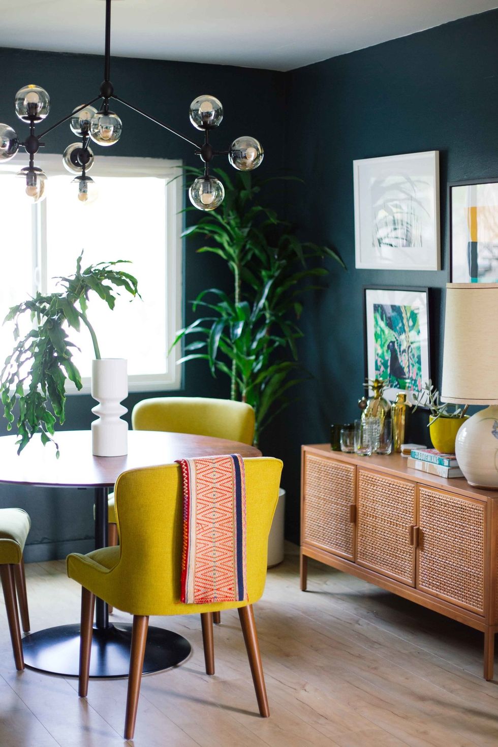 hình ảnh phòng ăn với tường sơn màu xanh mòng két, bàn ăn tròn, ghế vàng, đèn thả thủy tinh, tủ gỗ, tranh treo tường, cây xanh trang trí