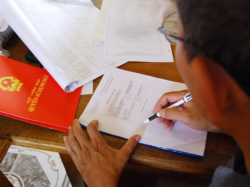 hình ảnh cận cảnh một người đàn ông đang viết trên giấy tờ, cạnh đó là cuốn sổ đỏ