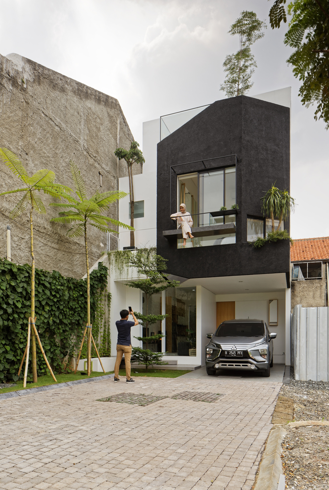 hình ảnh toàn cảnh mặt tiền ngôi nhà 2 tầng ở Indonesia với mảng tường sơn màu xám đen, khung cửa sổ kinh có cô gái ngồi vắt vẻo, tầng trệt có ô tô đang đậu và người đàn ông dùng điện thoại chụp ảnh căn nhà