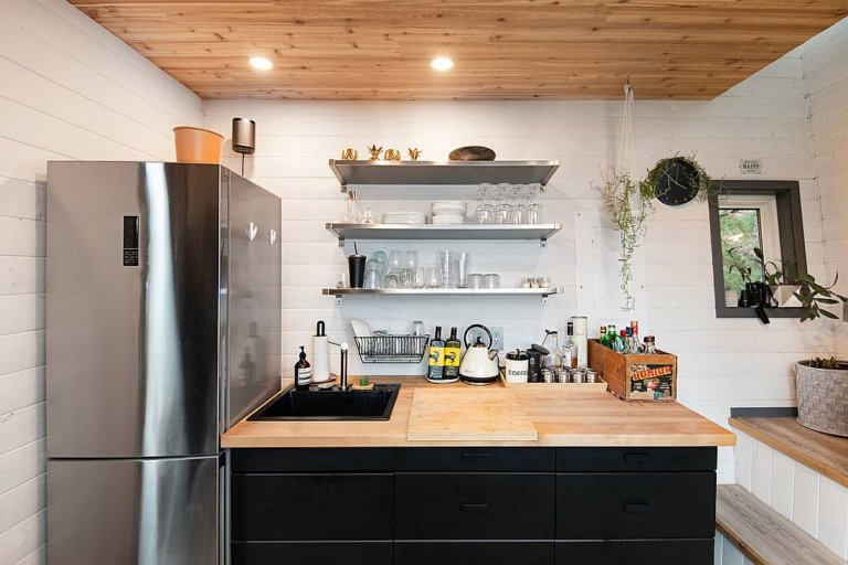 hình ảnh toàn cảnh phòng bếp với kệ mở gắn tường, bàn đảo tích hợp ngăn kéo màu đen, trần gỗ, đèn LED âm trần