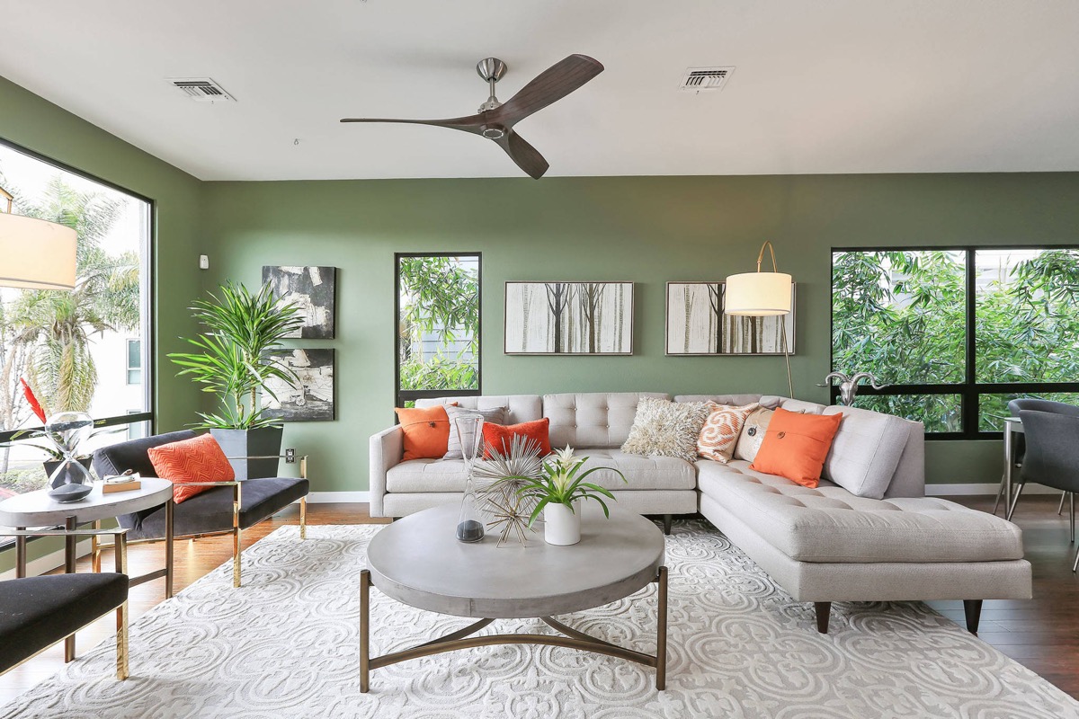 hình ảnh phòng khách màu xanh lá với sofa trắng xám, cửa sổ kính trong suốt, gối tựa màu cam bắt mắt