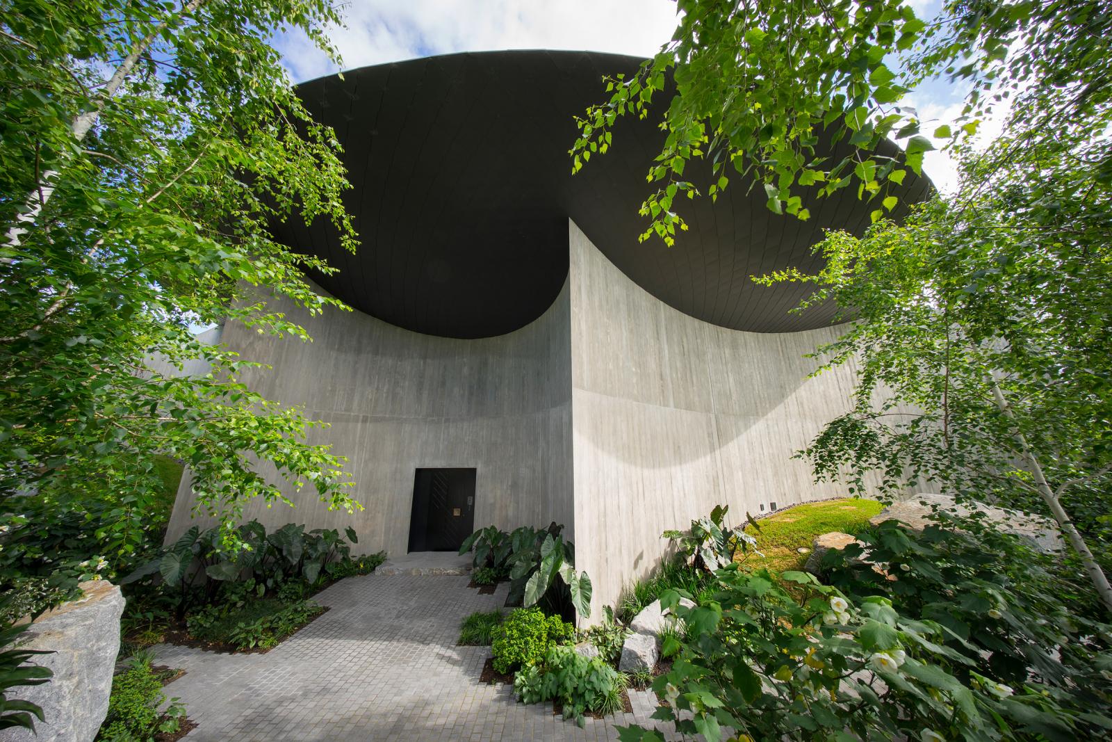 hình ảnh một góc nhà có mái hình đĩa ở Úc với tường bê tông uốn cong, vườn cây xanh tốt bao quanh