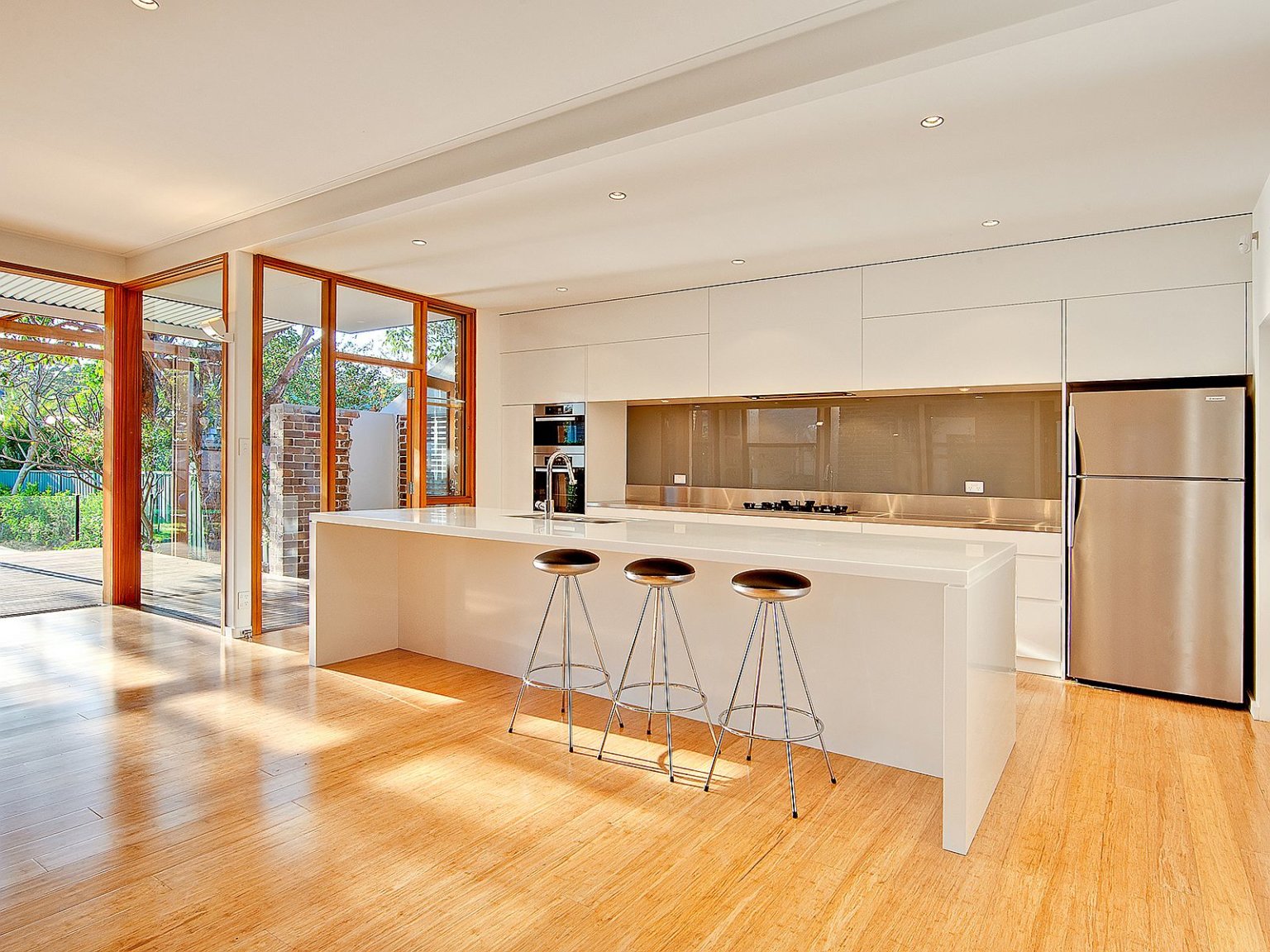 hình ảnh phòng bếp hiện đại với tông trắng chủ đạo cho trần, tường, tủ bếp, bàn bar, sàn gỗ sáng bóng nối dài ra sân hiên
