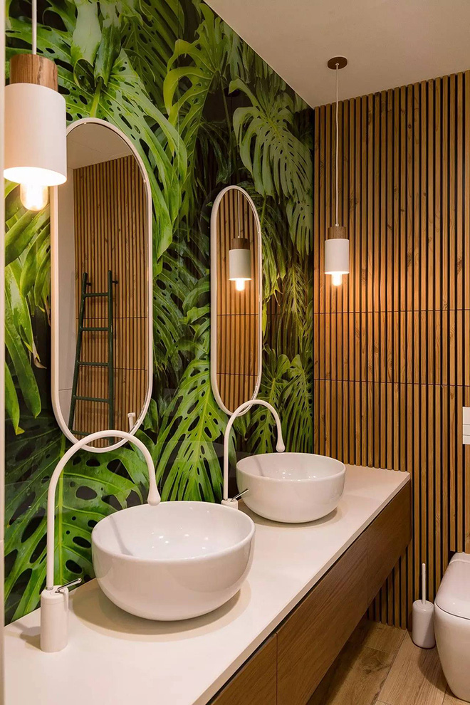 hình ảnh góc phòng tắm với bồn rửa đôi màu trắng, hai khung gương bầu dục treo tường, giấy dán tường họa tiết lá cây lớn
