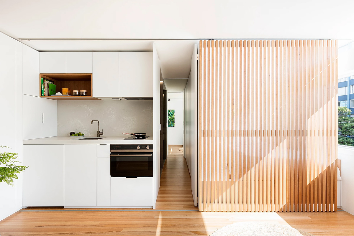 hình ảnh phòng bếp nhỏ với hệ tủ màu trắng cánh phẳng, cạnh đó là lam gỗ giấu gọn phòng ngủ