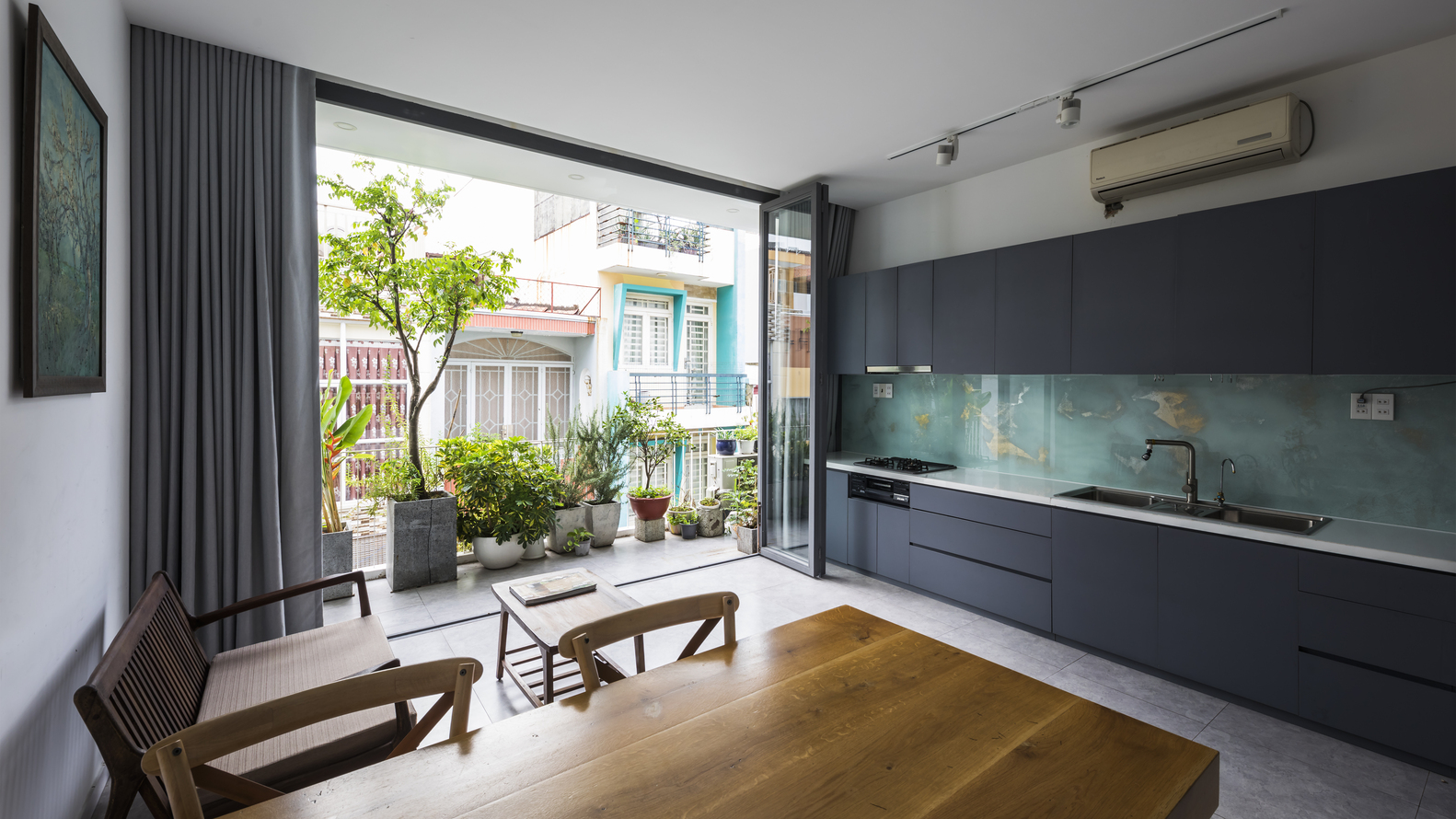 hình ảnh toàn cảnh phòng bếp ăn nhà phố 4 tầng với tủ bếp màu xám đen, bàn ăn bằng gỗ, cửa kính lớn mở ra khoảng xanh trồng cây
