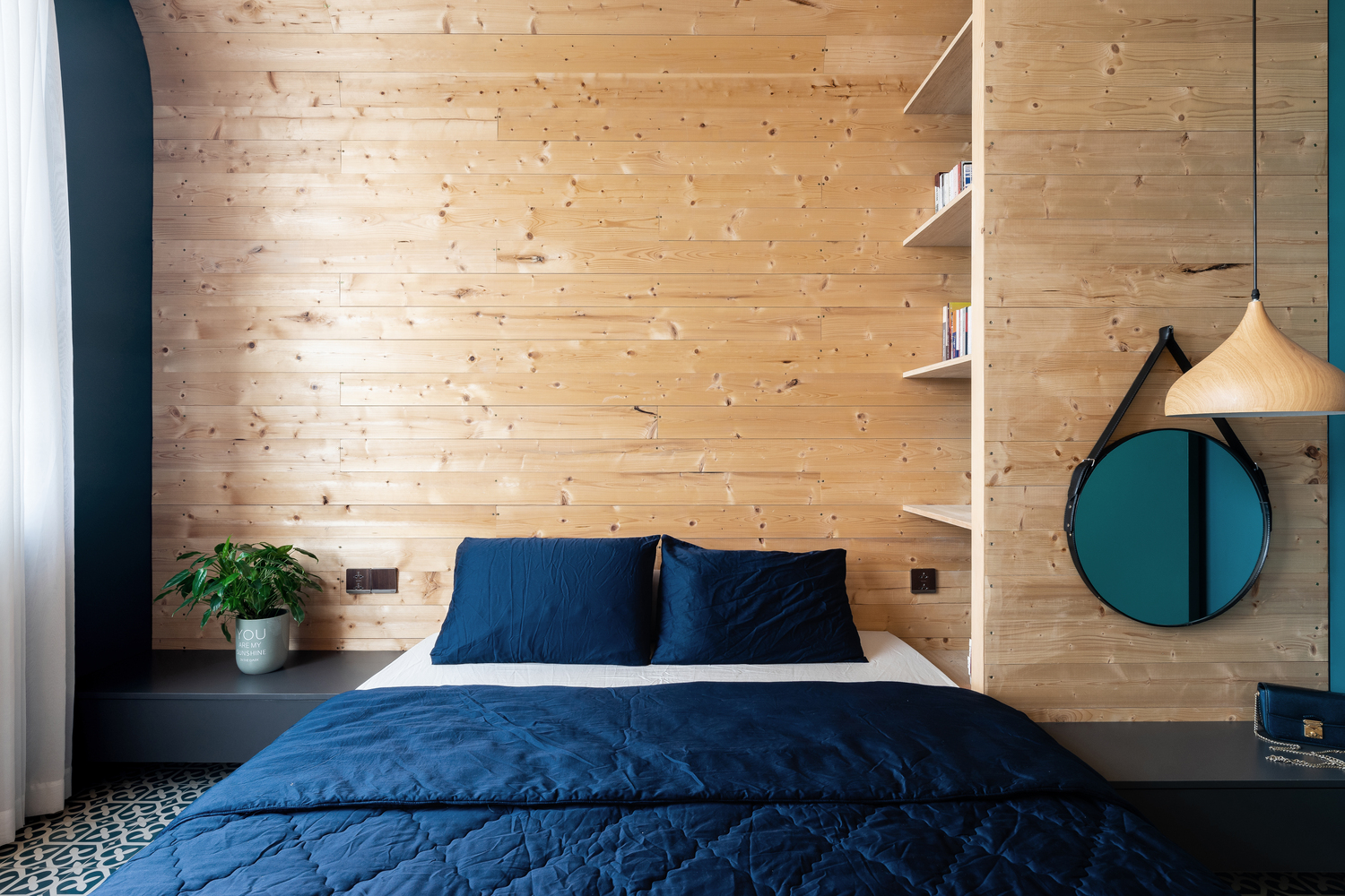 hình ảnh phòng ngủ nhỏ với tường đầu giường ốp gỗ, tủ kệ lưu trữ ngay cạnh, gương tròn trang trí, ga gối màu xanh nước biển