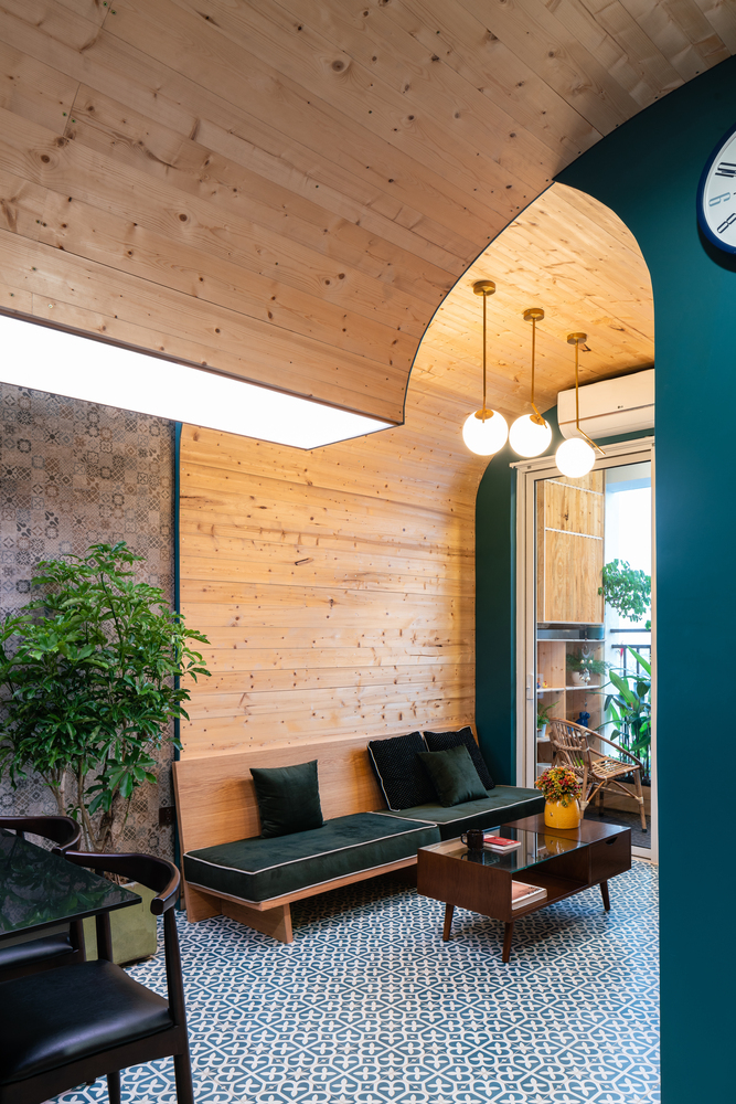 Hình ảnh phòng khách căn hộ nhỏ với tường và trần vòm ốp gỗ, sofa bọc nệm màu xanh dương, bàn trà kính, bộ ba đèn thả mặt dây chuyền ấm áp
