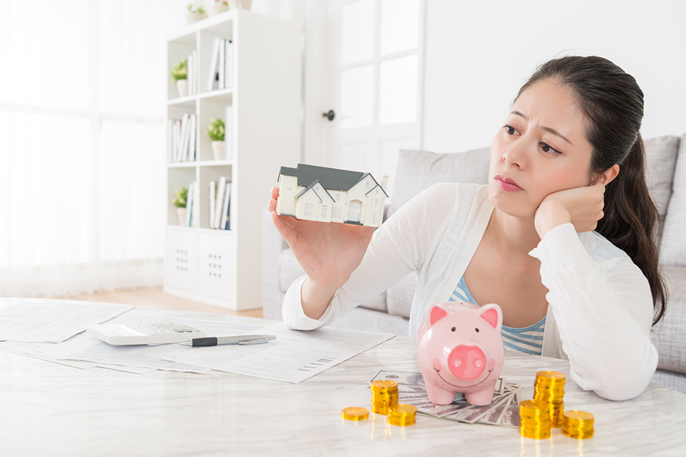 một người phụ nữ đang cầm tập tiền trên tay, mặt đăm chiêu suy nghĩ, trên bàn là mô hình ngôi nhà