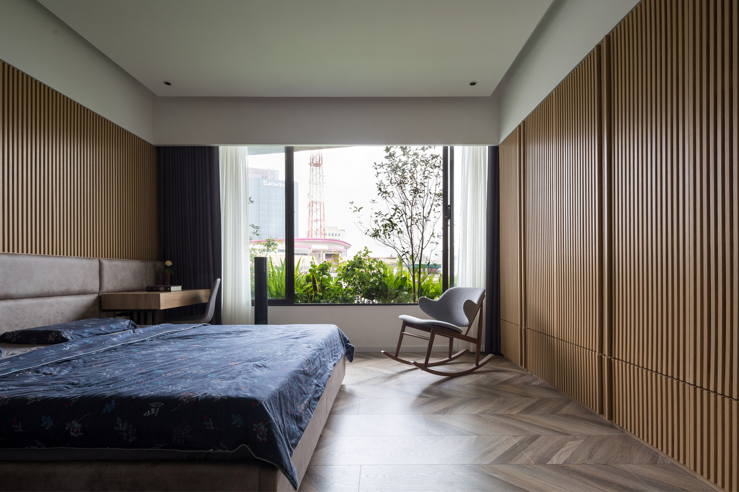 hình ảnh phòng ngủ với giường lớn, tường đầu giường ốp gỗ, ghế thư giãn ở góc phòng, cửa sổ kính lớn