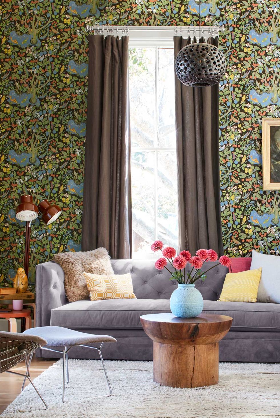 hình ảnh phòng khách nhỏ với sofa màu ghi xám, giấy dán tường họa tiết lá xanh, rèm cửa màu nâu lắp cao, đèn sàn, bình hoa tươi đặt trên bàn trà