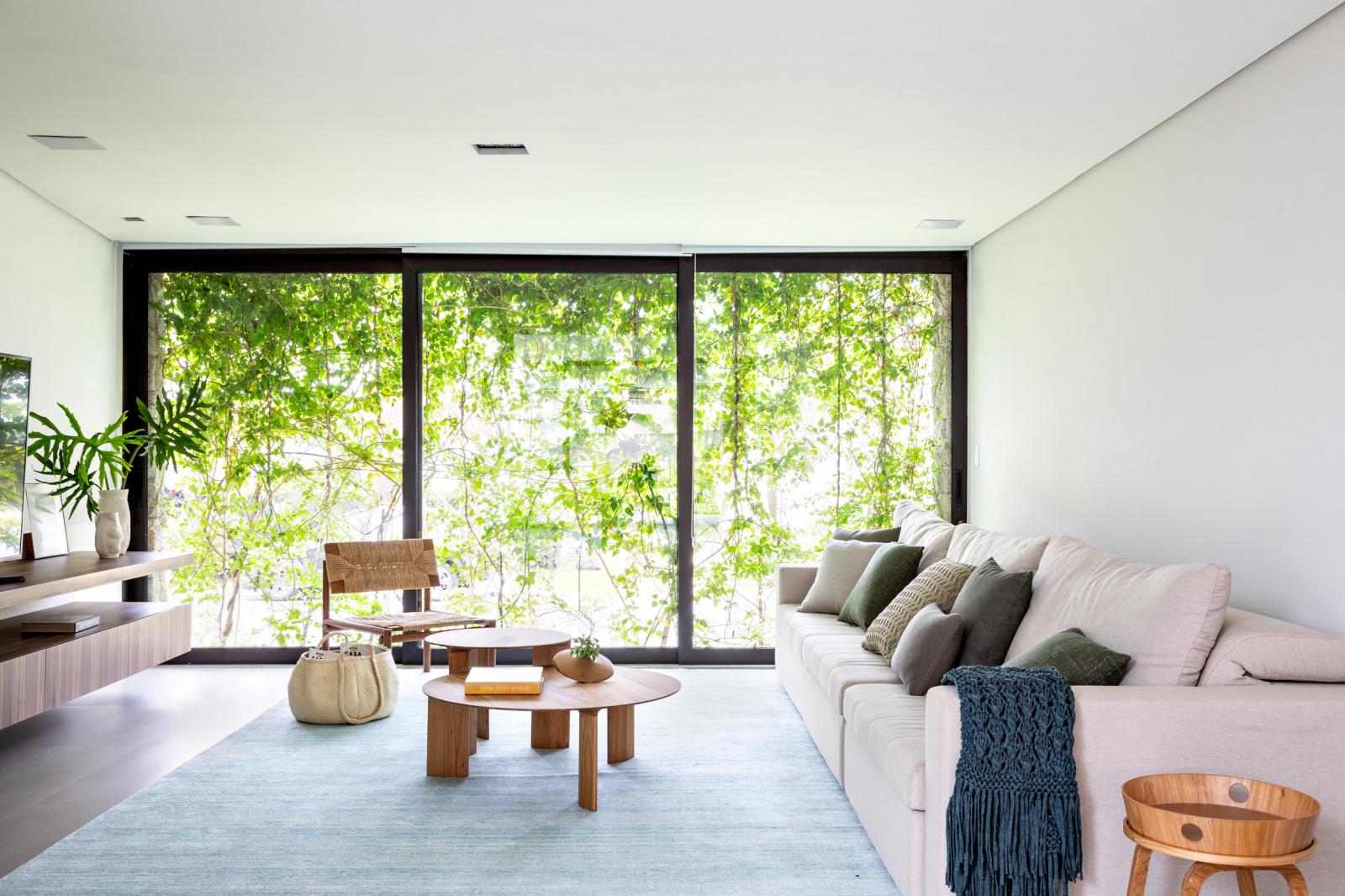 hình ảnh phòng khách ngập tràn ánh sáng với sofa trắng, tủ kệ tivi bằng gỗ, cửa kính trong suốt phản chiếu khung cảnh thiên nhiên xanh mát bên ngoài
