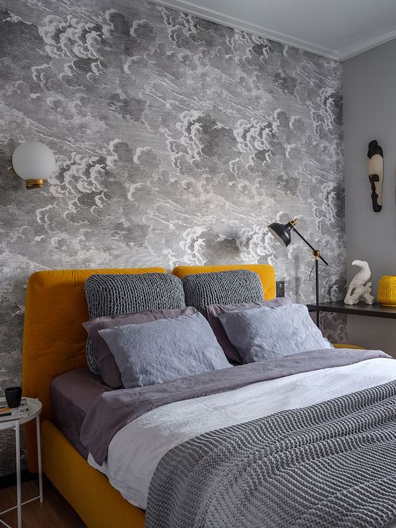 hình ảnh một phòng ngủ nhỏ với bức tường giấy dán hình đám mây màu xám, cùng tông ga gối, nổi bật trên khung giường màu vàng mù tạt.