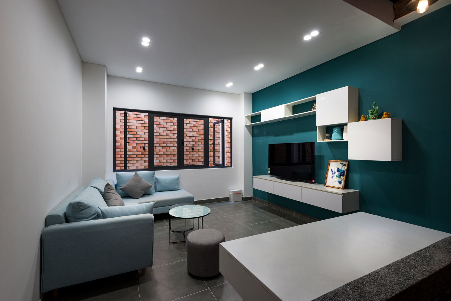 hình ảnh phòng khách nhà phố với sofa xám đặt đối diện với bức tường sơn màu xanh dương, cửa sổ kính mở ra tường gạch, khoảng thông tầng bên ngoài