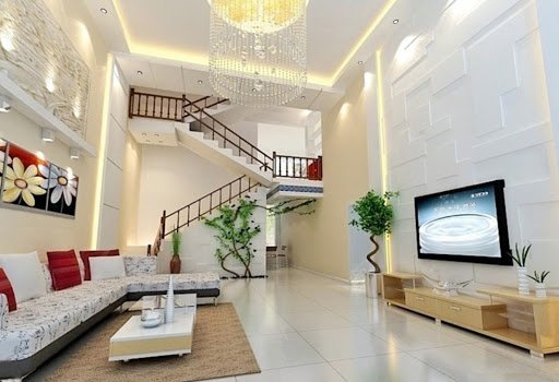 hình ảnh phòng khách nhà ống với trần caot hoáng, tường sơn màu trắng, sofa trắng, tranh hoa treo tường, đèn chùm lớn, cạnh đó là cầu thang lên tầng trên