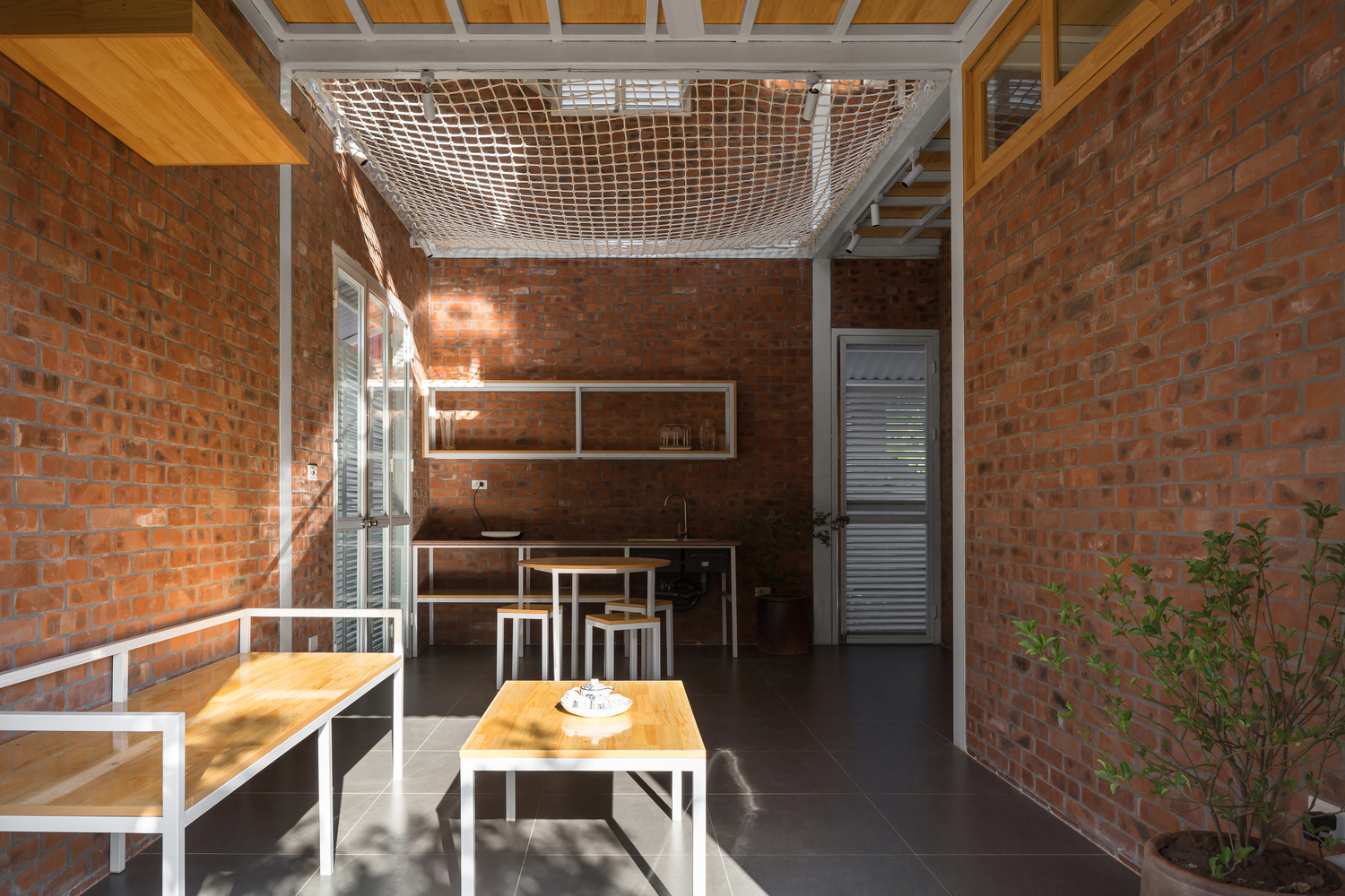 không gian bên trong nhà khung thép với tường gạch trần, lưới trắng, nội thất gỗ màu vàng sáng