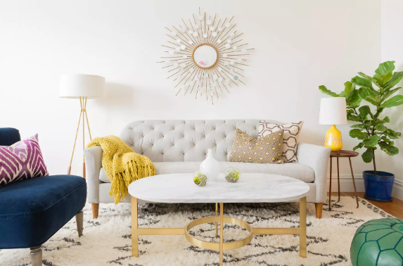 hình ảnh phòng khách với bàn trà cẩm thạch màu trắng, chân vàng đặt trên thảm, ghế sofa màu xanh coban, khăn mỏng màu vàng chanh, gương mặt trời treo tường, cây xanh 