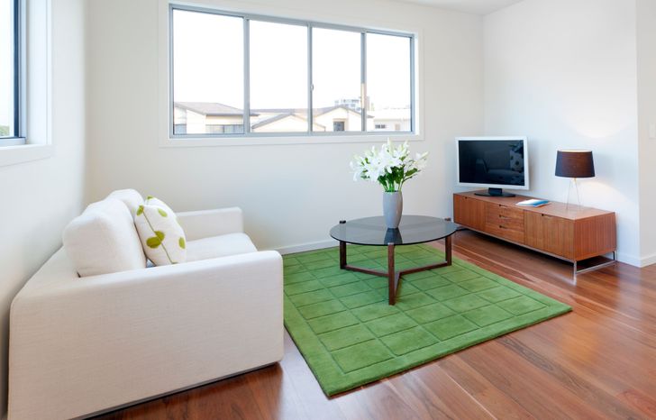 hình ảnh phòng khách với tường và trần sơn trắng, cửa sổ kính lớn, sofa trắng, bàn trà tròn, kệ tivi bằng gỗ, thảm trải sàn màu xanh lá cây