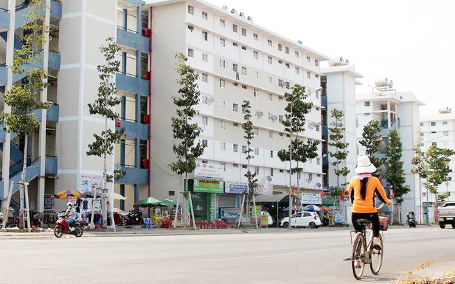 hình ảnh một tòa nhà ở xã hội màu trắng, xanh xám đã hoàn thiện và đi vào hoạt động, cô gái mặc áo vàng đạp xe trên đường lớn