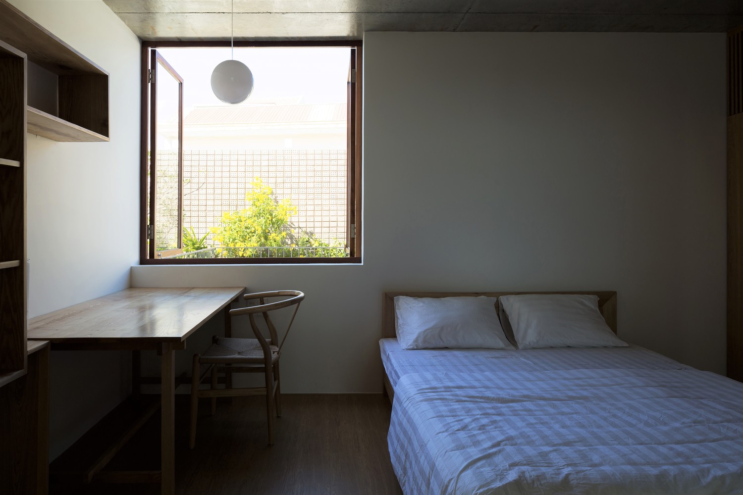 hình ảnh phòng ngủ nhỏ với giường nệm màu trắng, bàn gỗ, giá sách gỗ gắn tường, cửa sổ mở ra sân vườn