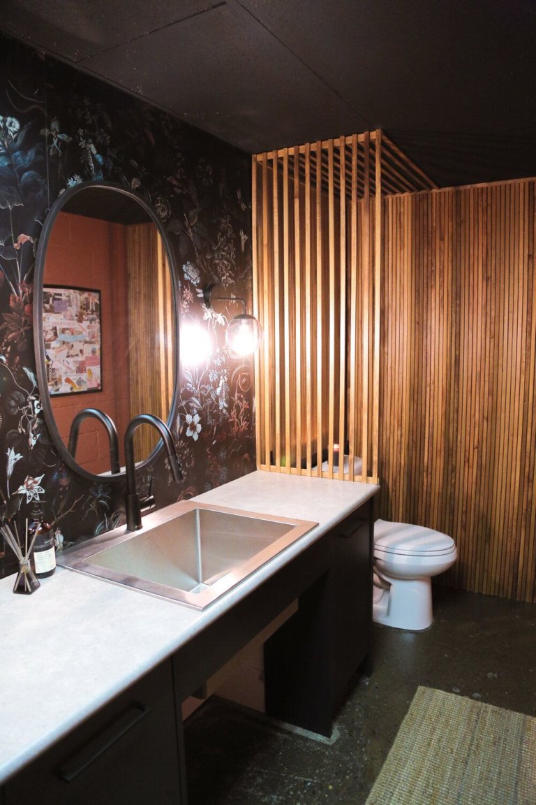 hình ảnh phòng tắm với giấy dán tường họa tiết hoa lá tối màu, gương hình elip, lam gỗ màu sáng