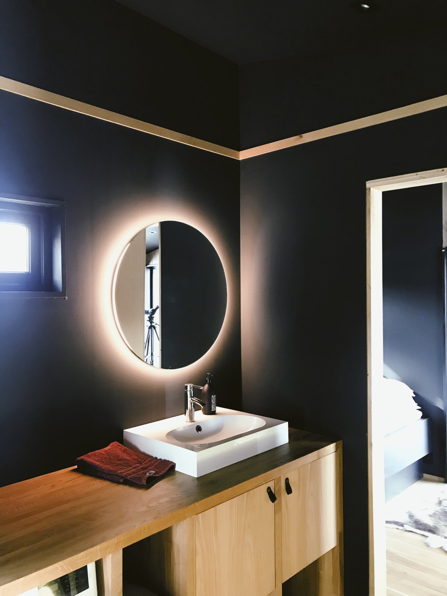 hình ảnh một góc phòng tắm với tường sơn màu xanh than, gương tròn kết hợp đèn LED, bồn rửa sứ trắng hình chữ nhật