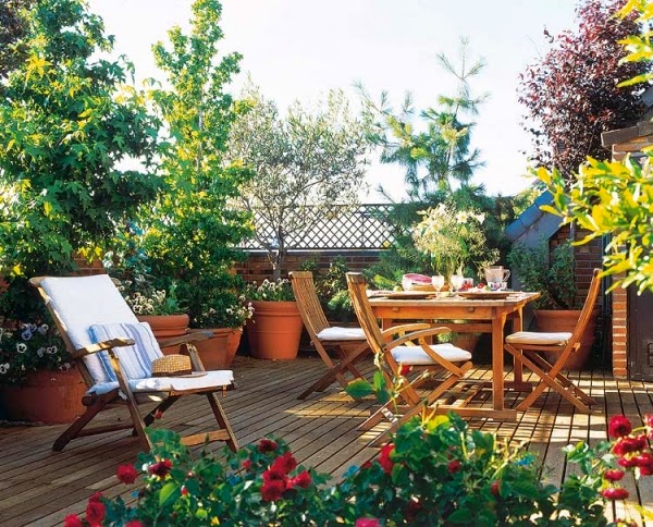 hình ảnh khu vườn trên sân thượng với sàn lát gỗ, bộ bàn ghế gỗ, xung quanh có nhiều chậu hoa màu đỏ, cây xanh tốt