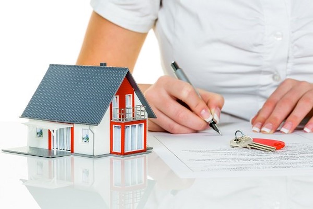 hình ảnh mô hình ngôi nhà, người mặc áo cộc tay màu trắng đặt bút ký vào giấy tờ minh họa cho việc sang tên sổ đỏ