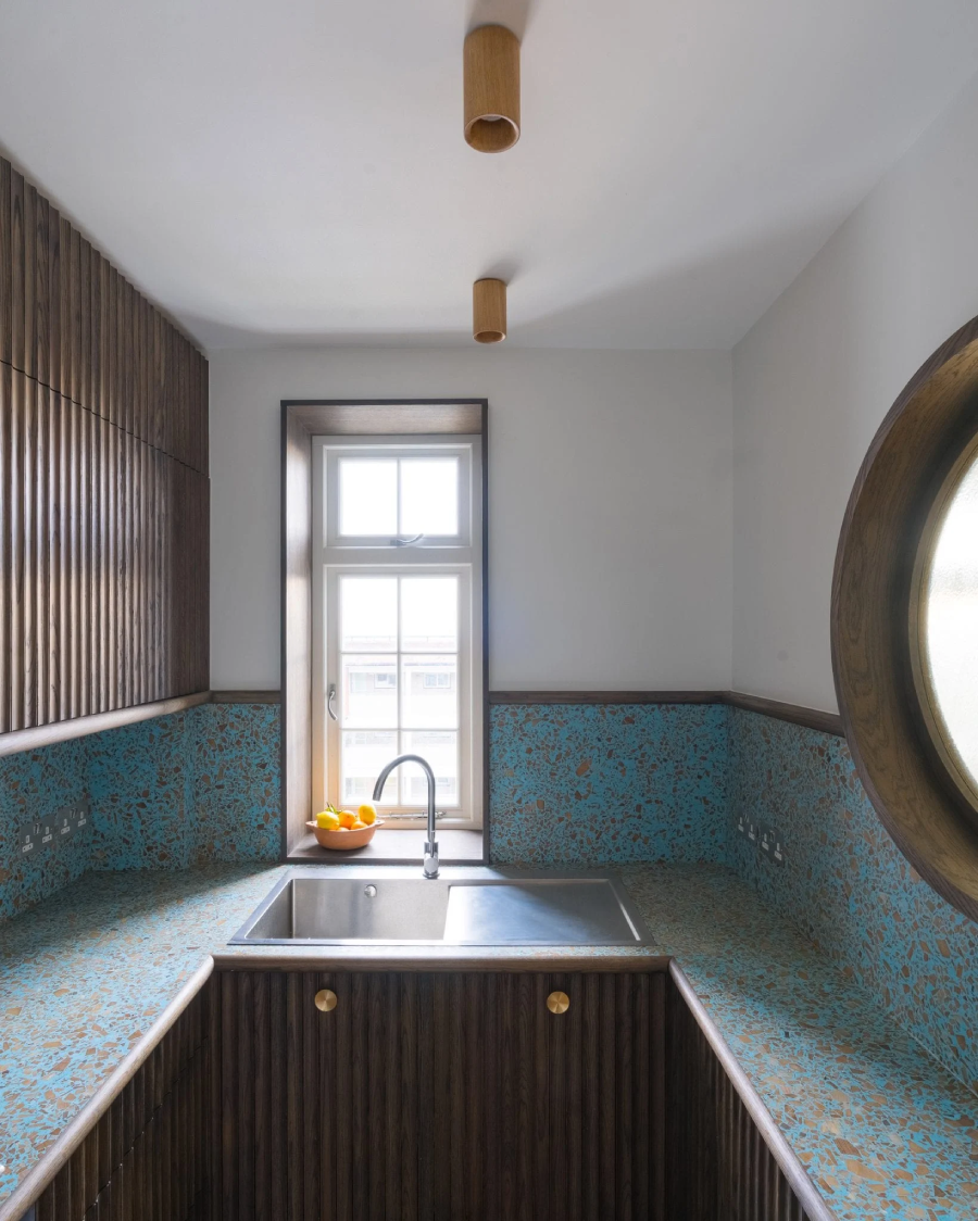 Trong nhà bếp nhỏ, bồn rửa đặt dưới một cửa sổ hẹp với mặt bàn màu xanh ngọc lam được hoàn thiện bởi một lớp vật liệu giống như đá mài.