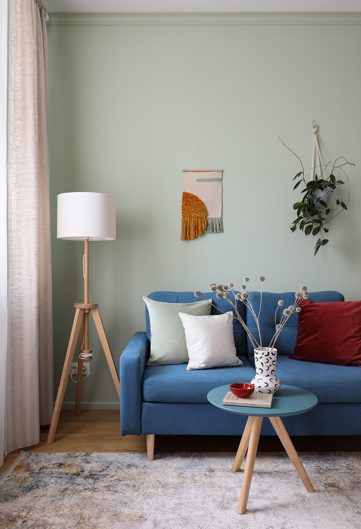 Góc phòng khách với sofa xanh, đèn sàn màu trắng, rèm cửa, chậu cây treo tường
